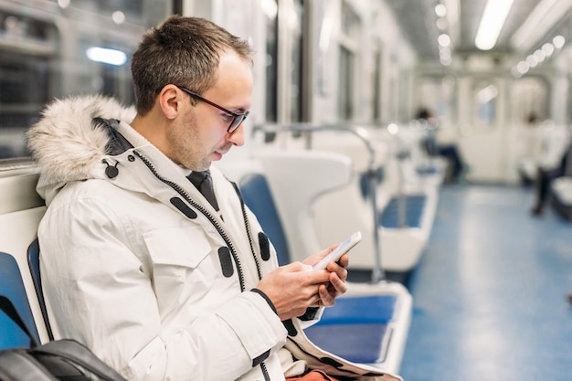 Heureux homme séduisant avec des lunettes en veste blanche monte dans le métro jusqu'à un rendez-vous Homme passe du temps dans un smartphone, il discute en surfant sur le Web ou en lisant des nouvelles Piège Internet Gros plan