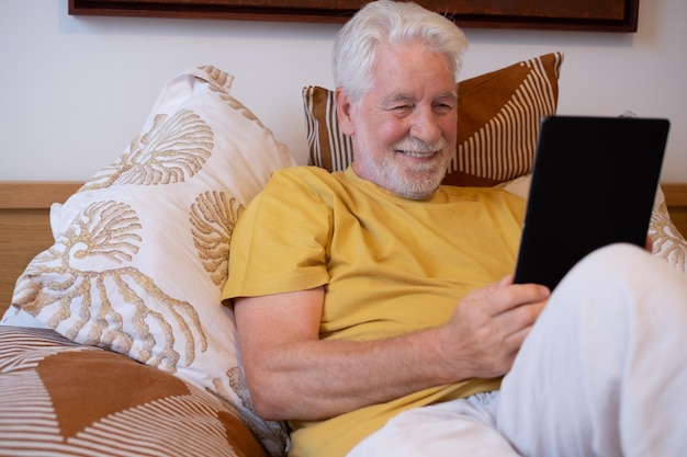 Heureux homme senior aux cheveux blancs assis sur le lit à la maison ou à l'hôtel en regardant une tablette numérique