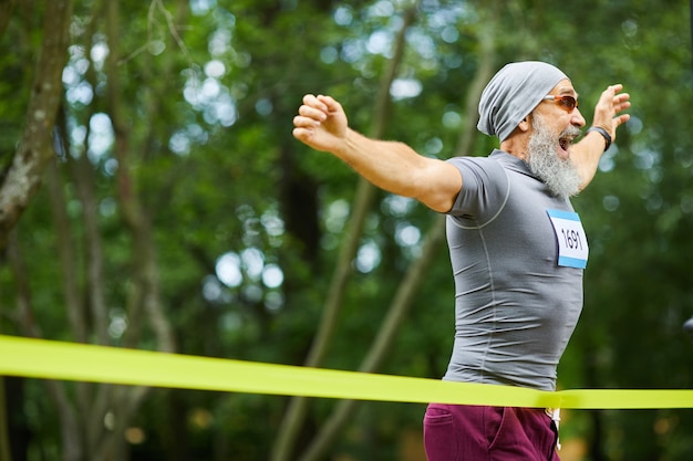 Heureux homme senior barbu portant casquette et lunettes de soleil remportant la première place dans la course de marathon, copiez l'espace