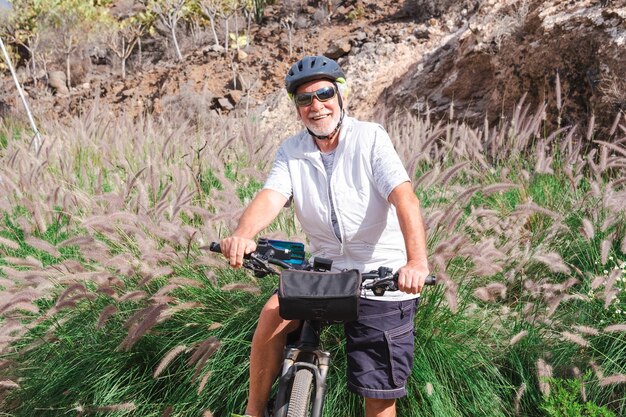 Heureux homme senior à cheval sur une route de campagne avec son vélo, cycliste portant un casque de sport et des lunettes de soleil