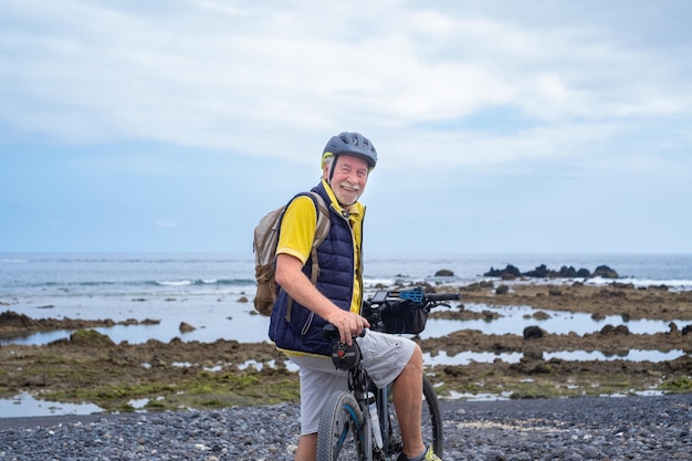 Heureux homme senior cycliste barbu le long de la plage de la mer portant un sac à dos portant un casque en cours d'exécution avec un vélo électrique regardant la caméra