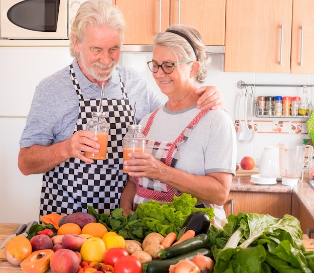 Heureux homme senior et sa femme souriant et buvant ensemble le jus de fruit qui vient d'être fait. Table en bois avec un grand groupe de fruits et légumes colorés. Alimentation équilibrée