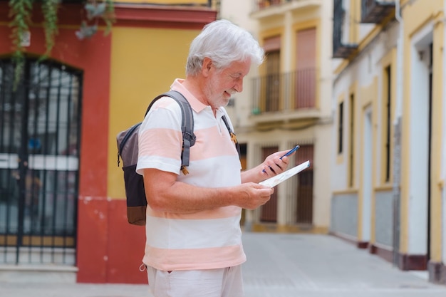 Heureux homme senior touriste tenant un sac à dos sur l'épaule marchant dans les ruelles de la vieille ville de Séville en Espagne tout en suivant la direction de la carte sur son téléphone portable