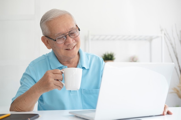 Heureux homme senior travaillant sur ordinateur portable et tenant une tasse de café à son domicile.