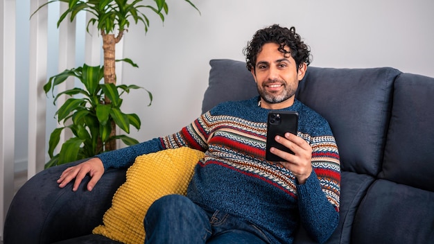 Heureux homme souriant à l'aide d'un smartphone assis sur un canapé à la maison. Homme mûr allongé sur un canapé, lisant des messages sur téléphone mobile. Espagnol caucasien se détendre à la maison.