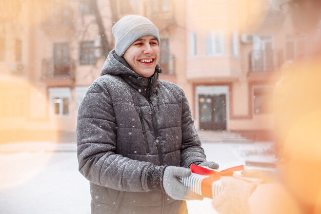 Heureux homme souriant reçoit un cadeau de sa petite amie pour la Saint-Valentin dans un parc d'hiver enneigé. Un jeune homme reçoit un cadeau à l'extérieur par une froide journée d'hiver. Concept de vacances et de cadeaux.