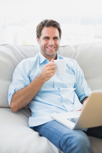 Heureux homme utilisant un ordinateur portable assis sur le canapé avec un café