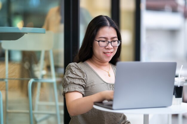 Heureux des indépendants asiatiques d'affaires femme décontractée travaillant avec un ordinateur portable avec une tasse à café et un smartphone dans un café comme l'arrière-plan, concept de communication