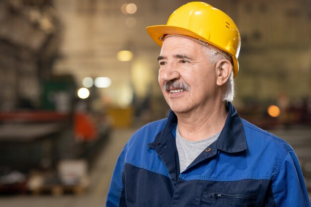 Heureux ingénieur mature ou contremaître en casque et vêtements de travail debout à l'intérieur d'une grande usine ou usine industrielle