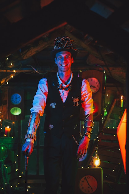 Heureux inventeur masculin en costume steampunk, chapeau haut de forme, lunettes et sourires de canne dans un atelier d'horlogerie