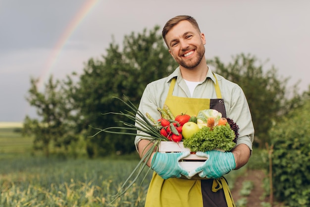 Heureux jardinier homme tenant un panier avec des légumes frais sur arc-en-ciel et jardin concept de jardinage de fond