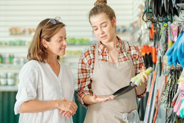Heureux jeune assistant de jardinage en tablier et chemise montrant de petites pelles à une cliente brune mature dans un grand supermarché