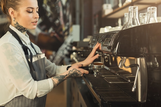 Heureux jeune barista tatoué faisant du café dans une machine à café professionnelle. Femme préparant une boisson. Concept de brassage de café pour les petites entreprises et les professionnels