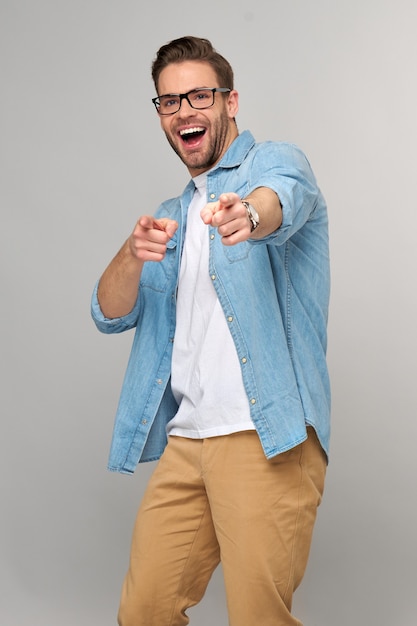 Heureux jeune bel homme en chemise de jeans pointant sur la caméra debout contre le mur gris