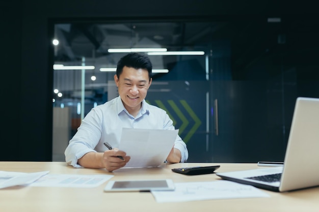 Heureux jeune comptable asiatique assis à un bureau dans le bureau tenant des documents dans ses mains