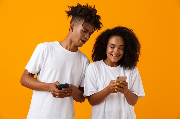 Heureux jeune couple africain mignon isolé sur un espace jaune à l'aide de téléphones mobiles.