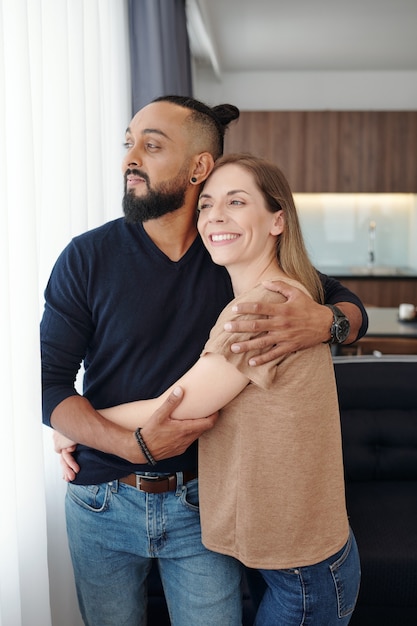 Heureux jeune couple amoureux s'embrassant et regardant par la fenêtre de l'appartement