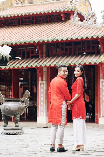 Heureux jeune couple asiatique en robes traditionnelles marchant vers le temple du bouddhisme, faisant demi-tour et souriant à la caméra