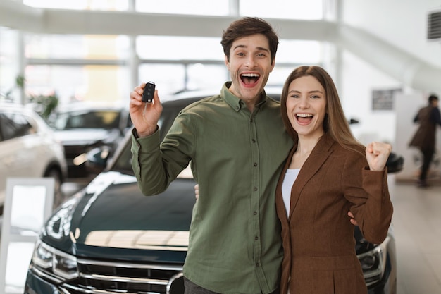 Heureux jeune couple caucasien faisant un geste oui montrant la clé de voiture achetant une nouvelle auto chez le concessionnaire