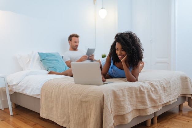 Heureux jeune couple détendu à la maison couché dans son lit sur la tablette et l'ordinateur