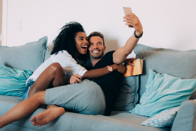 Heureux Jeune Couple Détendu à La Maison Dans Le Canapé En Lisant Un Livre Et En Regardant Le Téléphone Mobile