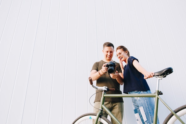 Heureux jeune couple élégant, homme et femme debout avec vélo, semble excitant tout en regardant des photos dans l'appareil photo après la prise de vue, à l'extérieur.