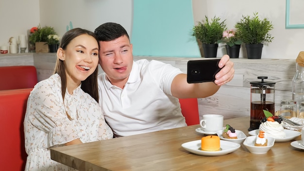 Heureux jeune couple fille guy fait selfie sur smartphone moderne assis à servi avec des gâteaux et des boissons table au café