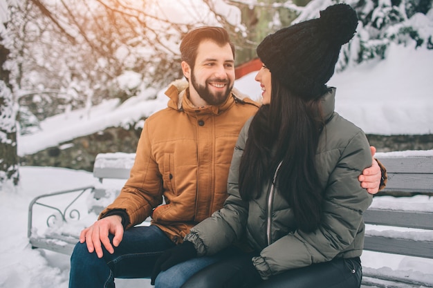 Heureux jeune couple en hiver. Famille à l'extérieur. homme et femme regardant vers le haut et riant. Amour, plaisir, saison et gens - marcher dans le parc d'hiver. Il neige, ils s'enlacent. Asseyez-vous sur le banc.