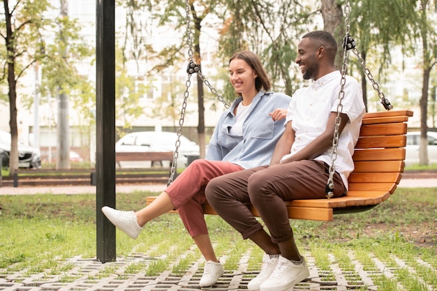 Heureux jeune couple interculturel en tenue décontractée assis sur une balançoire en bois dans un parc ou une place, discutant et riant tout en profitant de la journée d'été