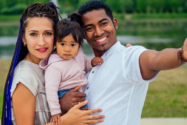Heureux jeune couple interracial transnational de race mixte passant du temps avec leur fille à l'aide d'un smartphone faisant un portrait de caméra photo selfie