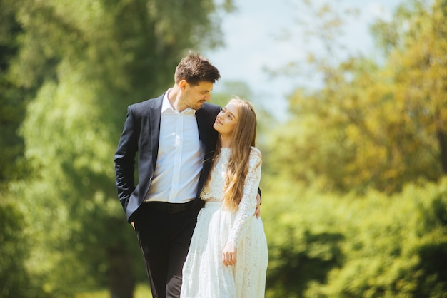 Heureux jeune couple de mariage en plein air dans le parc en se regardant, en marchant et en souriant au soleil.