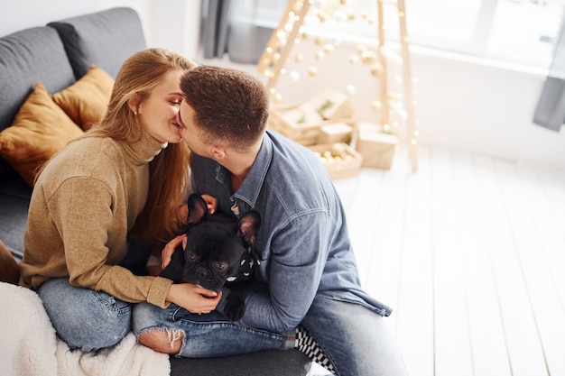 Heureux jeune couple moderne assis sur le canapé à la maison avec un arbre de Noël avec leur chien mignon.