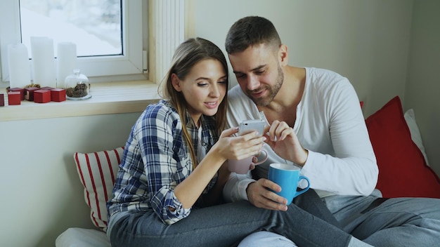 Heureux jeune couple parlant et parcourant les médias sociaux sur smartphone tout en étant assis sur le lit