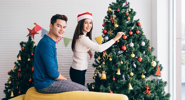 Heureux jeune couple portant des chandails s'amusant à décorer diverses boules sur le sapin de Noël dans le salon à la maison Profiter et célébrer les vacances de Noël