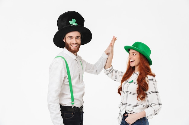 Heureux jeune couple portant des costumes, célébrant le jour de la Saint-Patrick isolé sur un mur blanc, s'amusant ensemble, high five