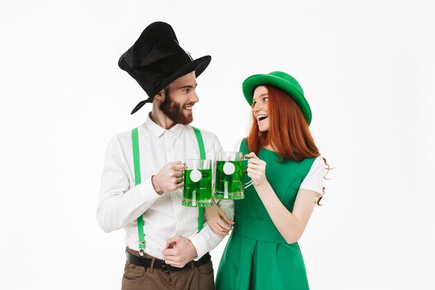 Heureux jeune couple portant des costumes, célébrant la Saint-Patrick isolé sur mur blanc, boire de la bière