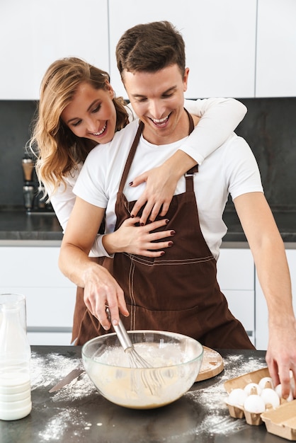 Heureux jeune couple portant des tabliers debout au bureau de la cuisine, faire de la pâte, s'étreindre