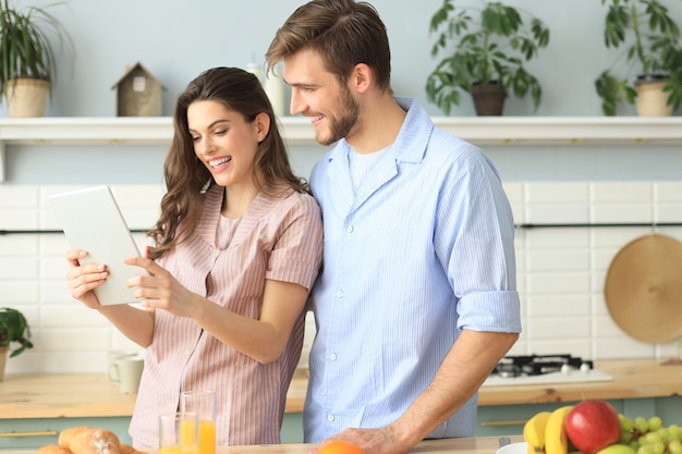 Heureux jeune couple en pyjama regardant du contenu en ligne sur une tablette et souriant tout en cuisinant dans la cuisine à la maison.