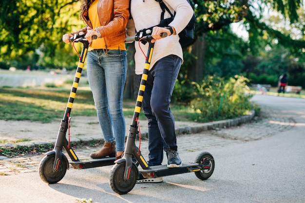 Heureux jeune couple s'amusant ensemble tout en conduisant des scooters électriques dans le parc de la ville.