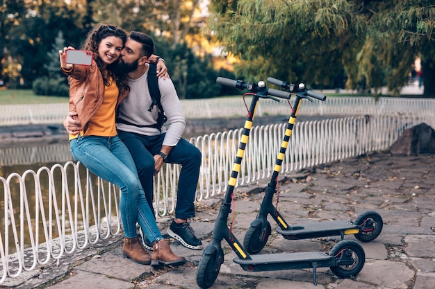 Heureux jeune couple avec des scooters électriques s'amusant ensemble dans le parc de la ville et prenant une photo de selfie.