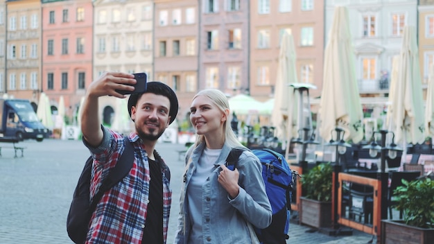 Heureux jeune couple de touristes faisant selfie sur smartphone dans le centre-ville. Ils ont des sacs de touristes.