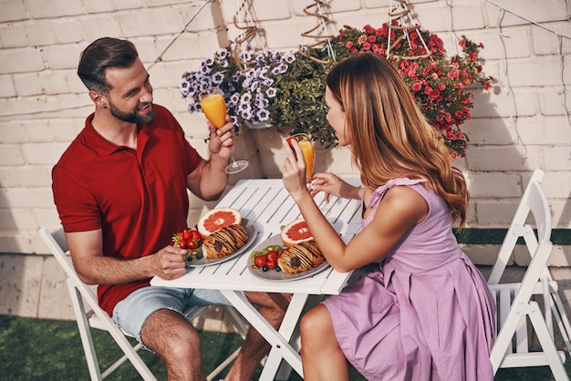 Heureux jeune couple en vêtements décontractés prenant son petit déjeuner et souriant assis sur la terrasse sur le toit à l'extérieur