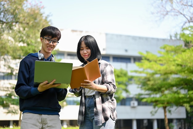 Heureux jeune étudiant asiatique féminin et masculin se tient avec un ordinateur portable et un livre scolaire