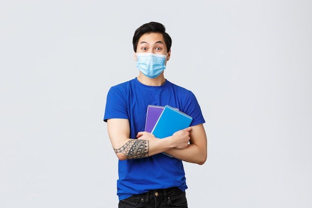 Heureux jeune étudiant asiatique mâle heureux en masque médical rubrique campus, souriant et tenant des cahiers