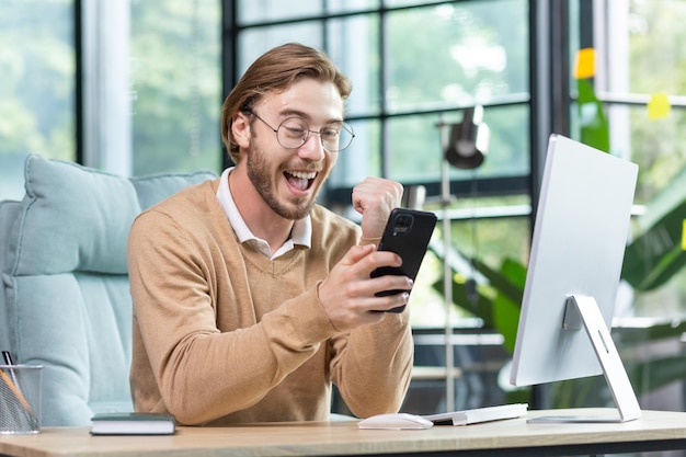 Heureux jeune étudiant à lunettes assis au bureau à la maison devant l'ordinateur tenant le téléphone à la main