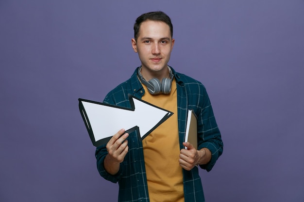 Heureux jeune étudiant de sexe masculin portant des écouteurs autour du cou tenant un bloc-notes sous le bras et une flèche pointant vers le côté regardant la caméra isolée sur fond violet