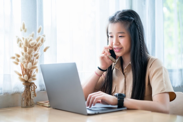 Photo heureux de jeune étudiante adolescente asiatique en train de parler en utilisant un smartphone occasionnel en utilisant un ordinateur portable