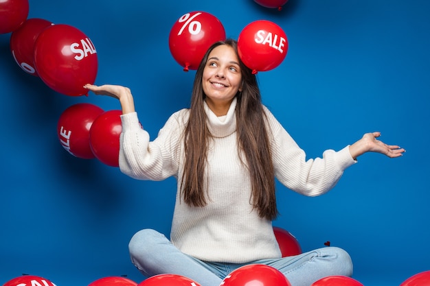 Heureux jeune femme assise et à la recherche de ballons à air rouge en vente, isolé sur mur bleu