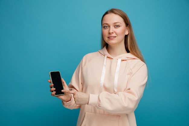 Heureux jeune femme blonde tenant et pointant le doigt sur téléphone mobile