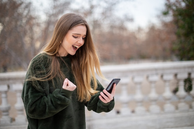 Heureux jeune femme criant et en gesticulant avec la main tout en se tenant dans le parc et en regardant smartphone moderne